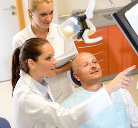 A middle-aged man looking at a screen while the dentist and dental assistant explain what is showing