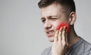 Man wearing a grey tee is holding his hand to his cheek with a look of pain on his face